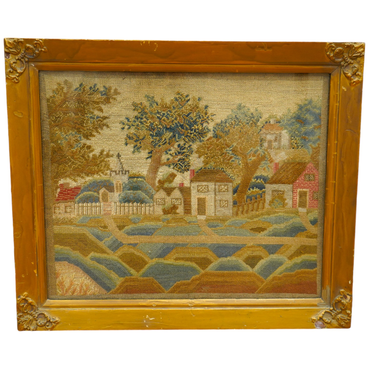 A framed 19th century woolwork of a village scene of cottages, houses, a church amongst trees and a picket fence with paths leading to another, within an undulating foreground, the leaves of the trees worked in various s